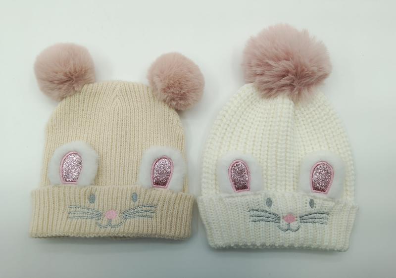 Two knit baby beanies in cream and white with glitter bunny ear patches, embroidered whiskers, and pink faux fur pompoms on top - BSCI + Oeko-Tex + GRS certified