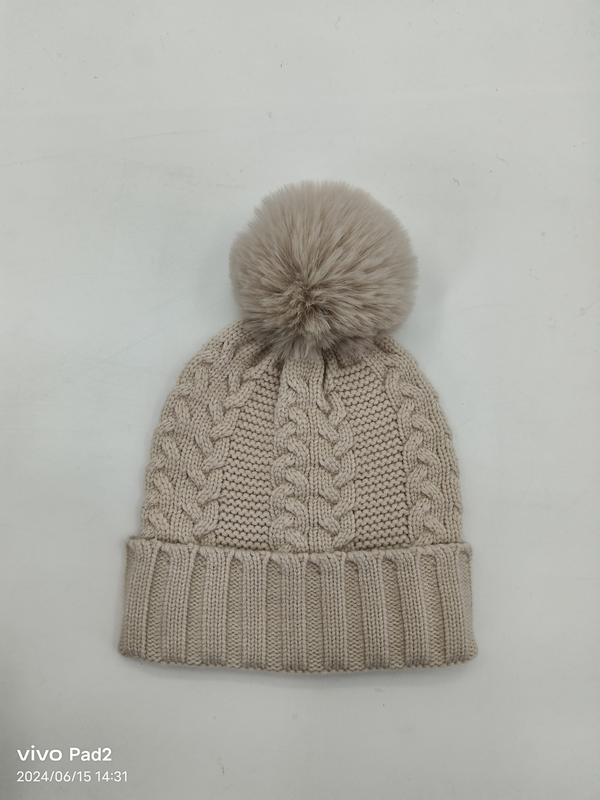 Beige cable knit winter beanie hat with large faux fur pom pom and ribbed cuff, laid flat on white background - EU REACH compliant material