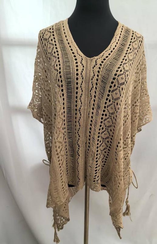 Beige open-weave crochet poncho top with floral lace pattern, V-neckline, short sleeves, and tassel fringe hem on mannequin - tiered pricing available ask quote