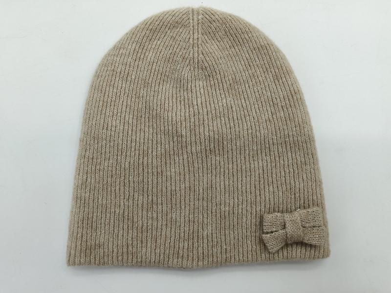 Beige ribbed knit beanie hat with small knitted bow accent on the side, laid flat on white background - Oeko-Tex Standard 100 certified