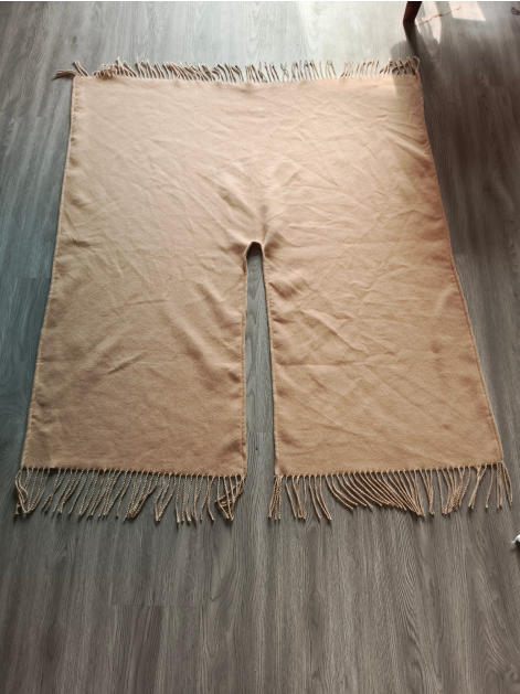 Flat lay of a beige woven poncho with center neck slit and twisted fringe along top and bottom edges on wood floor - tiered pricing available ask quote