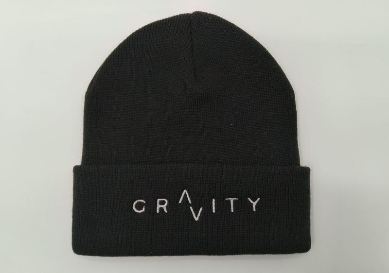 Black ribbed knit cuffed beanie hat with white embroidered GRAVITY logo on folded cuff, laid flat on white background - EU REACH compliant material