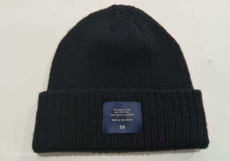 Black ribbed knit beanie hat with folded cuff and blue woven brand label patch on white background - low MOQ 50pcs trial order