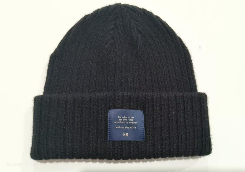Black ribbed knit winter beanie hat with folded cuff and blue woven label patch on white background - bulk order 20-day delivery
