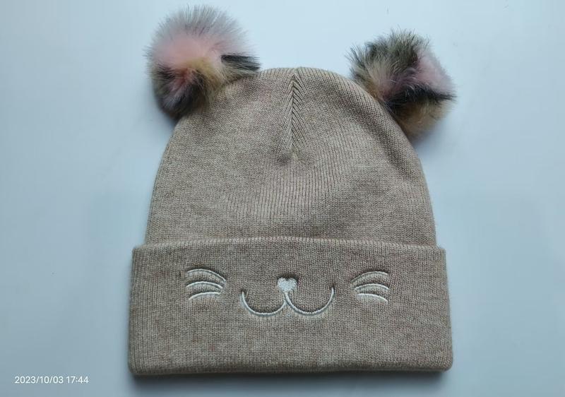Beige knitted children's beanie hat with two multicolor fur pom poms and embroidered cat face design on cuff - wholesale price for USA buyers
