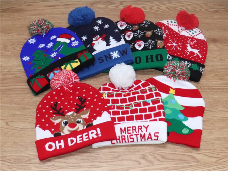 Six colorful Christmas knit beanies with pom-poms featuring reindeer, Santa, snowman, and holiday tree patterns on wooden surface - custom packaging design free