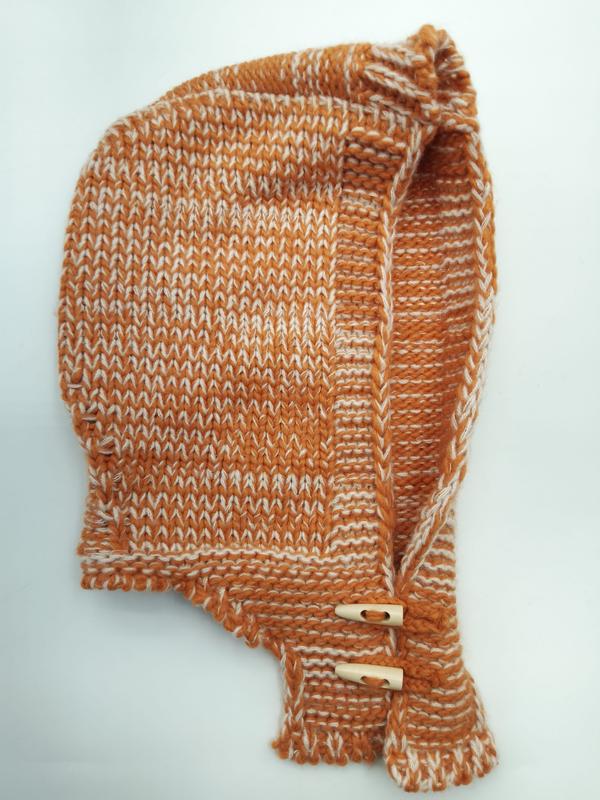 Hand-knitted baby bonnet in burnt orange and cream marled yarn with wooden toggle buttons on chin straps, laid flat on white background - express production 15 days