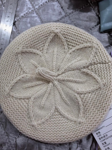 Cream knitted beret hat with raised floral leaf cable pattern at crown, laid flat on silver fabric - bulk order 500+ pcs extra 8% off