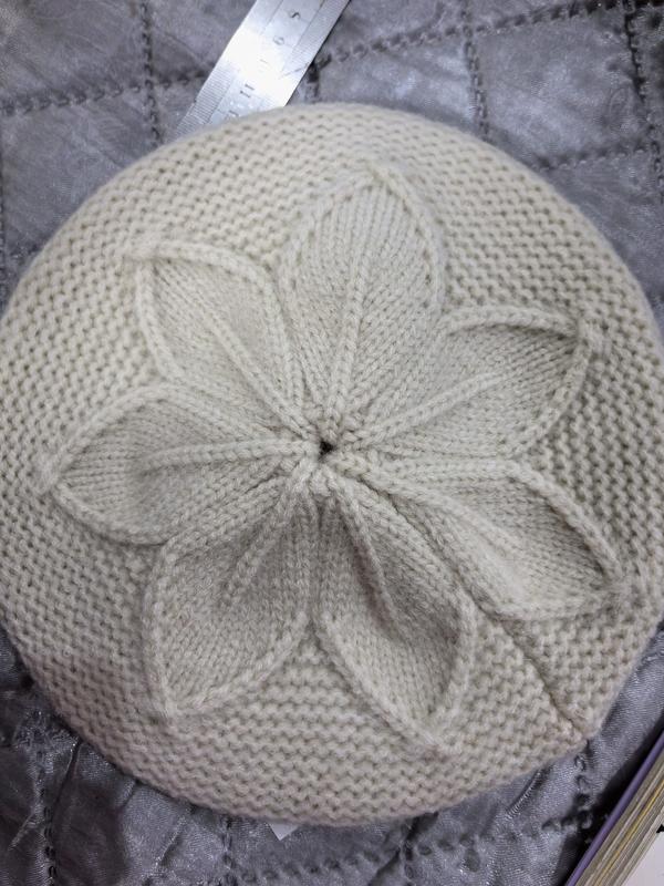 Top view of a cream knitted beret with a raised petal flower cable pattern at the crown, laid flat on gray quilted fabric - factory direct price no middleman
