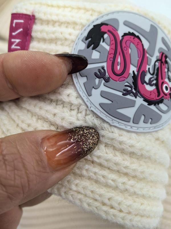 Close-up of cream chunky knit winter beanie hat held by hand with glitter ombre nails, featuring round pink snake rubber patch badge - jacquard custom pattern accepted