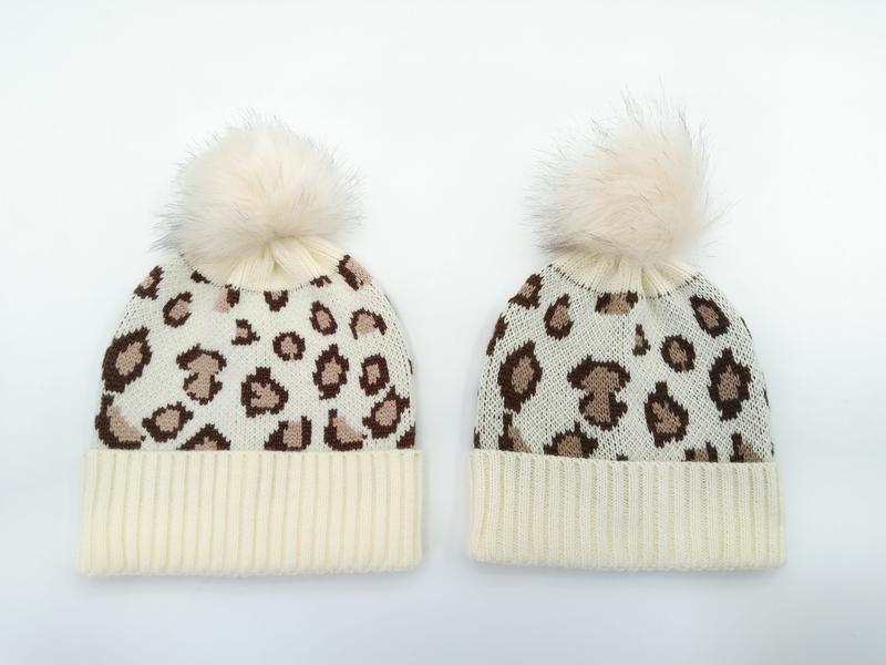 Two cream ivory leopard print knit beanies with fluffy faux fur pom-poms, adult and child sizes on white background - hotel amenity scarf supplier