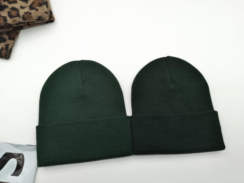 Two dark forest green ribbed knit cuffed beanie hats laid flat on white surface next to leopard print fabric - wholesale price for Europe buyers