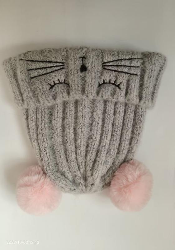 Grey ribbed knit winter beanie hat with embroidered cat face, whiskers, nose, and two pink fluffy pom poms at the base - wholesale price for Middle East buyers