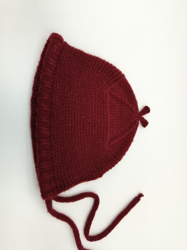 Hand-knitted dark red baby bonnet with garter stitch texture, ruffle edge, chin tie strings, and pointed top, laid flat on white background - WhatsApp quote in 2 hours