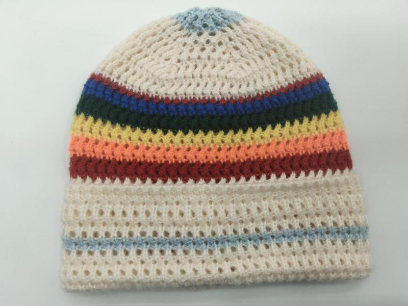 Handmade crochet beanie hat with rainbow multicolor stripes on cream open-weave background, laid flat on white surface - winter accessories wholesaler
