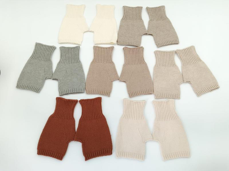 Seven pairs of handknit baby bloomers in cream, gray, taupe, beige, rust, blush, and sand laid flat on white background - hotel amenity scarf supplier