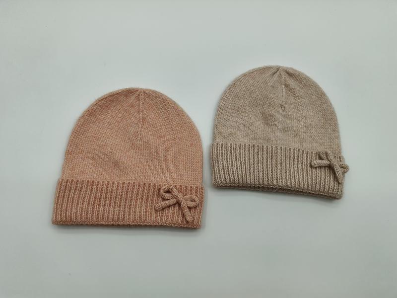 Two soft knit beanie hats with bow accents laid flat, one blush pink and one beige, ribbed cuff design - tiered pricing available ask quote