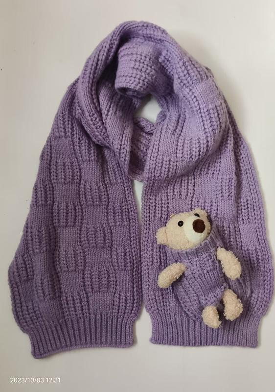 Purple lavender chunky knit scarf laid flat with a small cream teddy bear plush peeking out of a knitted pocket - gift shop wholesale supplier