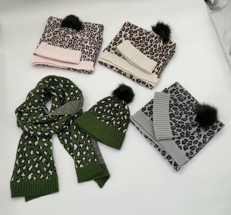 Four leopard print knitted hat and scarf sets in pink, cream, green, and gray with black faux fur pompoms displayed on white surface - ISO 9001 quality controlled