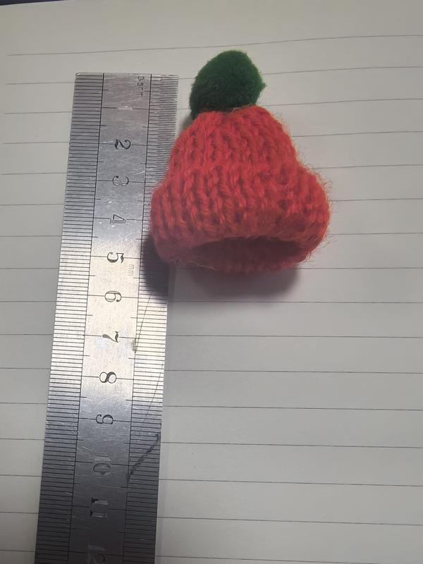 Tiny handknitted red wool beanie hat with green pom-pom measured against a ruler showing approximately 4cm height - winter accessories wholesaler
