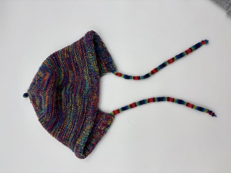 Handknit baby bonnet in multicolor variegated yarn with earflaps and braided tie strings, laid flat on white background - bulk order 500+ pcs extra 8% off