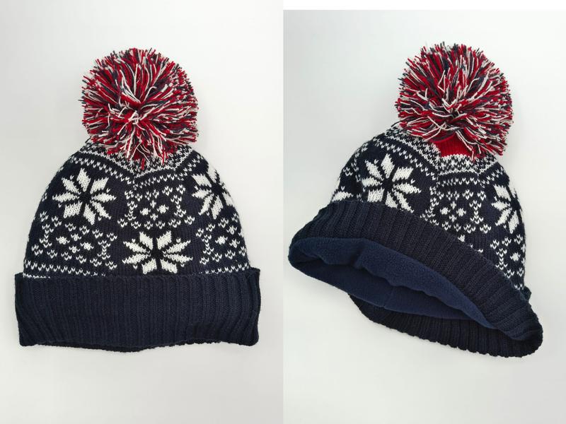 Navy blue knit beanie hat with white Fair Isle snowflake pattern, red and white multicolor pom-pom, and fleece-lined interior shown front and side view - bulk order 20-day delivery