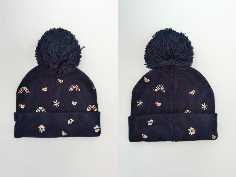 Navy blue knit beanie hat with large pom pom and colorful embroidered rainbows, hearts, stars and flower motifs, shown front and side view - jacquard custom pattern accepted