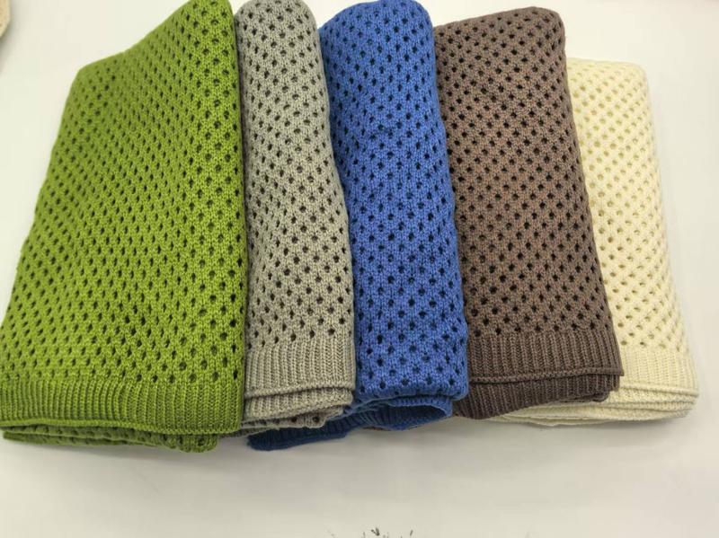 Five folded open-weave knit cotton throw blankets in green, grey, blue, brown, and cream stacked on white surface - Oeko-Tex Standard 100 certified