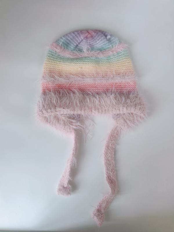 Handknit pastel rainbow striped baby beanie with fluffy pink eyelash yarn trim and long tie strings, laid flat on white surface - EU REACH compliant material