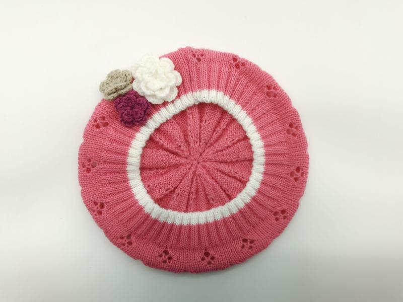 Pink knitted beret laid flat showing white circular stripe, eyelet lace pattern, and three small crochet flowers in white, mauve, and beige - EU REACH compliant material