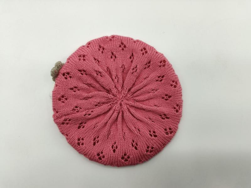 Flat lay of a pink knitted beret with radiating lace floral eyelets and a small tan pompom on a white background - ISO 9001 quality controlled