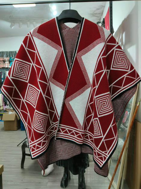 Red and white knitted poncho with bold geometric diamond and square patterns displayed on a hanger in a retail store - certified scarf manufacturer China - winter accessories wholesaler