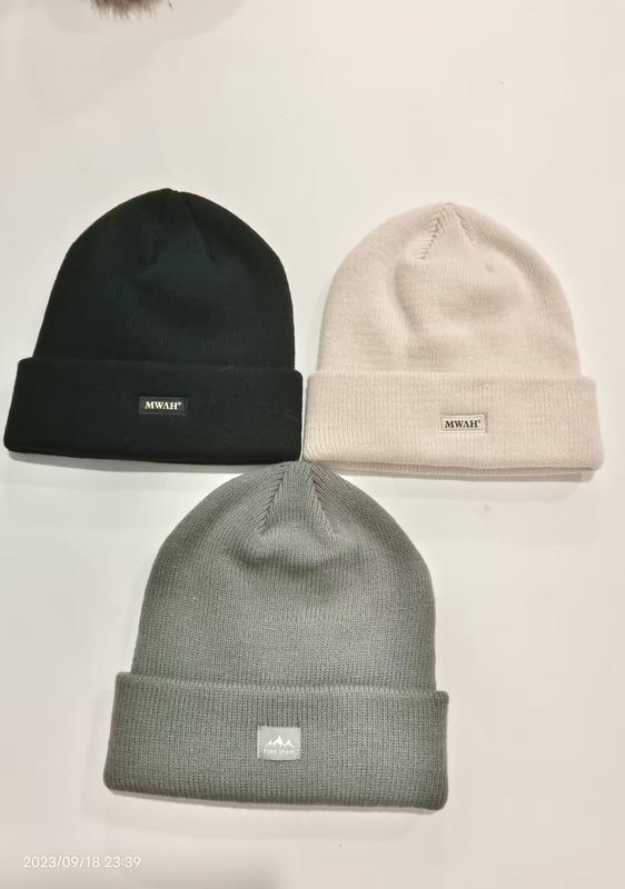 Three ribbed knit cuffed beanies laid flat in black, cream, and grey with small brand patch labels on white background - custom Pantone color matching