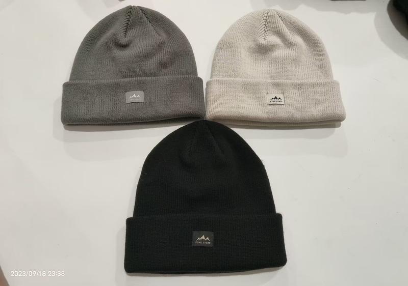 Three ribbed knit cuffed beanies in black, grey, and cream laid flat on white surface with mountain logo patch - custom packaging design free
