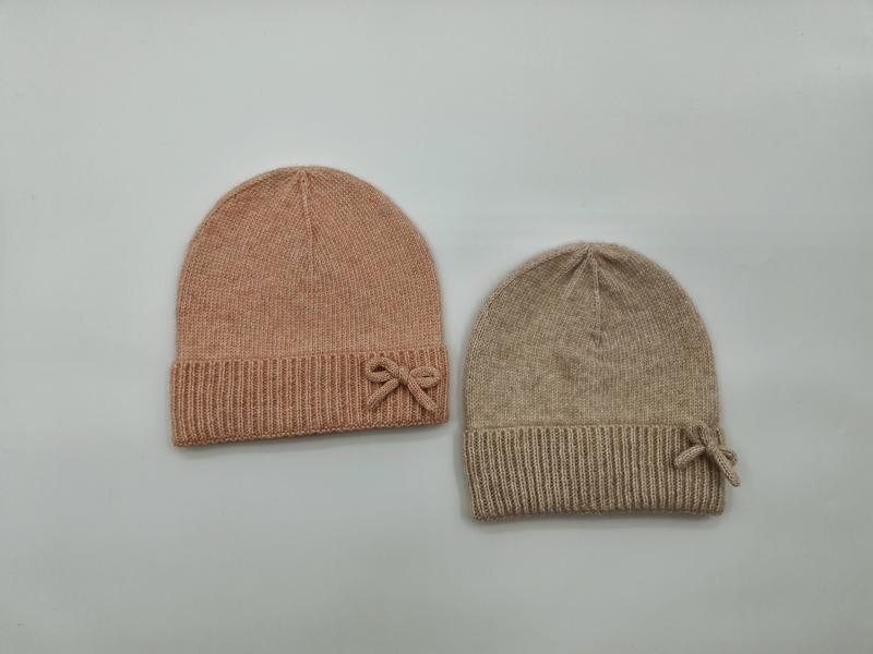 Two soft knit beanie hats with ribbed cuff and bow detail in blush pink and beige, laid flat on white background - wholesale price for Middle East buyers