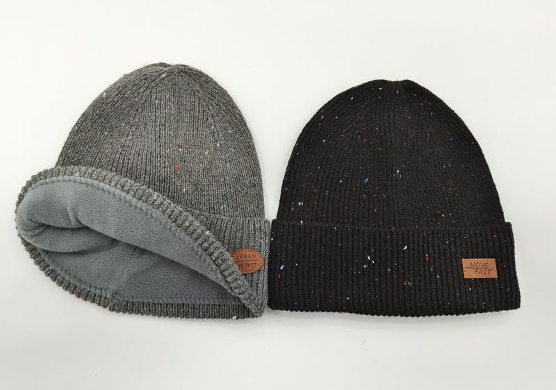 Two speckled knit winter beanies side by side: grey fleece-lined rolled beanie and black ribbed cuffed beanie with leather patch - ISO 9001 quality controlled